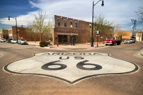 A view of a street in Winslow, Arizona, featuring a Route 66 sign and historic buildings in the background.