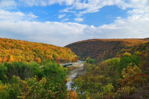 A scenic view of a river surrounded by autumn-colored hills under a partly cloudy sky.