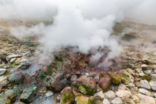 Steam rises from rocky terrain in a geothermal area, surrounded by colorful mineral deposits and misty clouds.