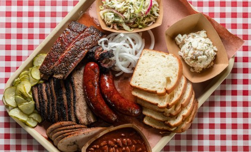 A platter of barbecue featuring ribs, sausage, brisket, pickles, onions, coleslaw, potato salad, and sliced bread.