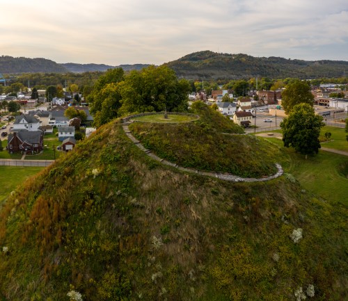Aerial view of a grassy hill with a circular path, surrounded by a small town and rolling hills in the background.