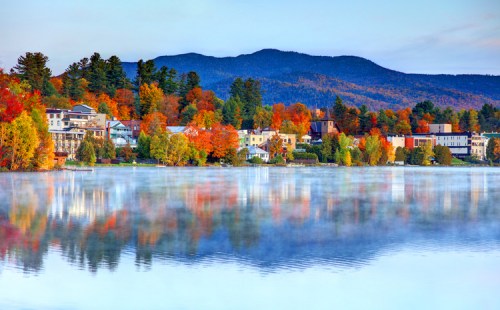 A serene lake reflects colorful autumn trees and a mountain backdrop, with a charming town along the shore.