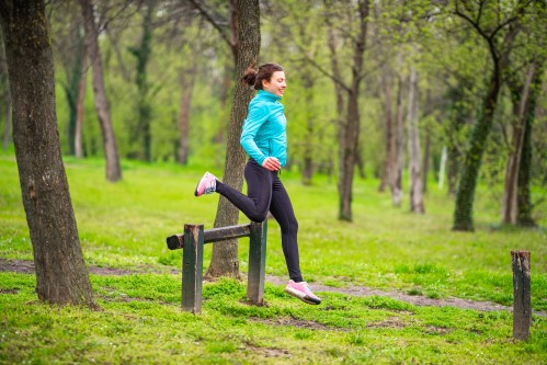 A woman in a blue jacket jumps over a wooden barrier in a green park surrounded by trees.