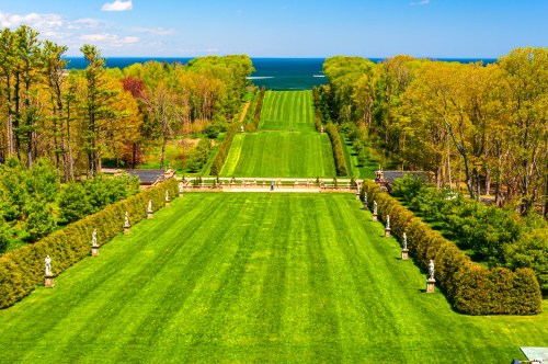 A lush green garden with neatly trimmed lawns, bordered by trees, leading to the ocean under a clear blue sky.