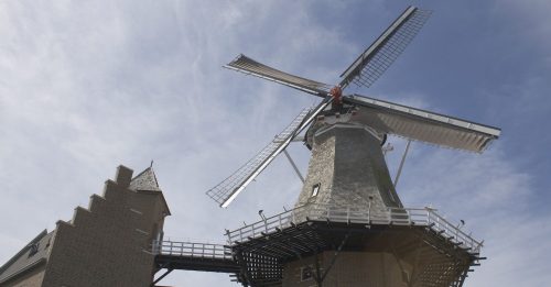 A tall windmill with large blades, set against a blue sky, featuring a brick base and a traditional architectural style.
