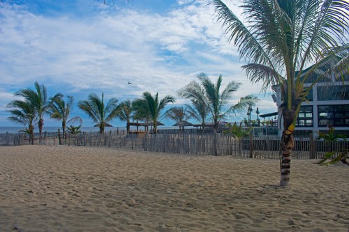 A sandy beach with palm trees, a wooden fence, and a restaurant in the background under a cloudy sky.