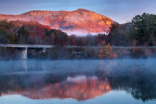 A serene landscape featuring a mountain reflected in a foggy lake, with autumn foliage and a bridge in the foreground.