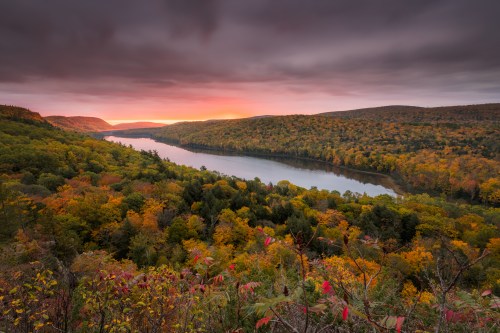 A scenic view of a river winding through vibrant autumn foliage under a dramatic sky at sunset.