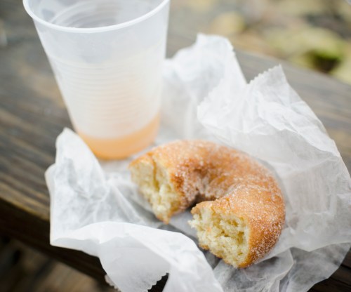 A partially eaten donut on wax paper next to a clear cup of beverage on a wooden surface.