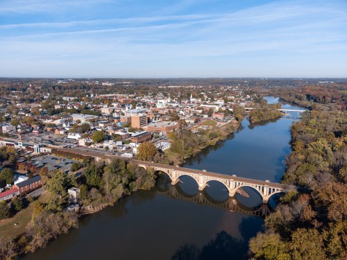 Aerial view of a town with a river and a bridge, surrounded by trees and buildings under a clear blue sky.