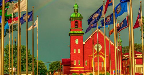 A red church with a green dome stands under a rainbow, surrounded by flags on a sunny, cloudy day.