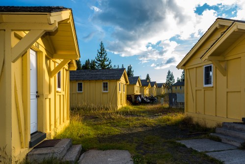 A row of yellow cabins under a blue sky with scattered clouds, surrounded by grass and trees.