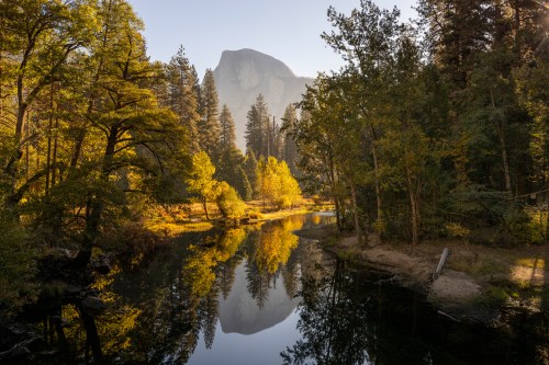 A serene landscape featuring a river reflecting trees and a mountain in the background, bathed in warm morning light.