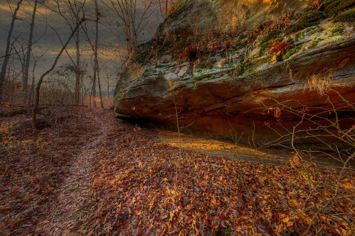 A rocky outcrop surrounded by autumn leaves and trees, with a winding path leading into the distance at sunset.