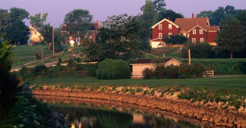 A serene landscape featuring a calm canal, lush greenery, and quaint houses under a soft morning light.