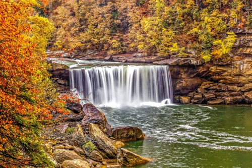 A serene waterfall cascades over rocks, surrounded by vibrant autumn foliage in warm hues of orange and yellow.