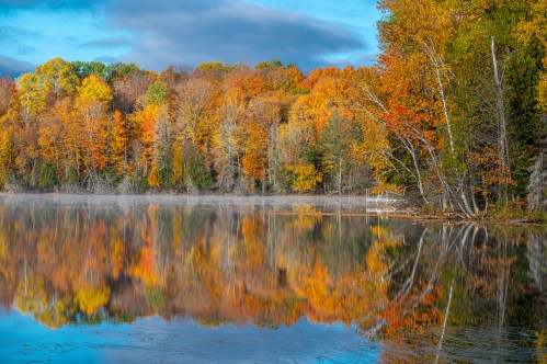 A serene lake reflects vibrant autumn foliage in shades of orange, yellow, and green under a clear blue sky.