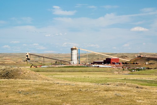 Industrial facility with silos and conveyor belts set against a clear blue sky and grassy landscape.