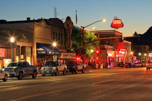 A street scene at dusk featuring shops, parked cars, and illuminated signs in a small town.