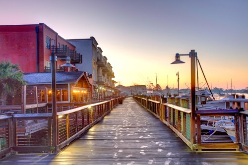 A wooden pier lined with buildings and boats, illuminated by a colorful sunrise.