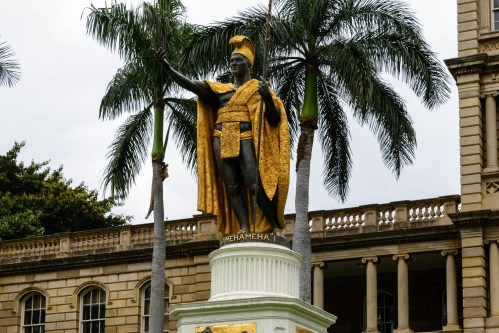 Statue of King Kamehameha I, adorned in gold, surrounded by palm trees in front of a historic building.
