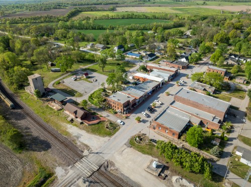 Aerial view of a small town with green fields, buildings, and a railway crossing.
