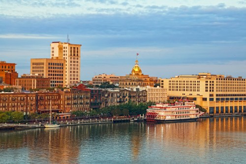 A scenic view of a city skyline along a river, featuring historic buildings and a riverboat at sunset.