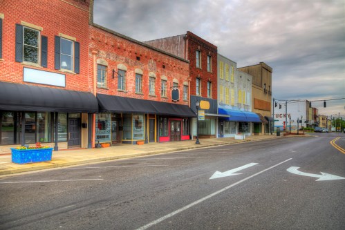 A quiet street scene featuring brick storefronts with awnings, under a cloudy sky in a small town.