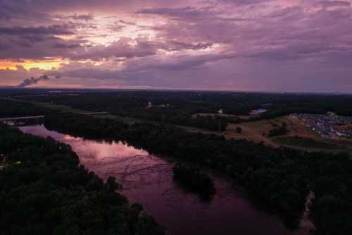 Aerial view of a river at sunset, surrounded by lush greenery and distant buildings under a cloudy sky.