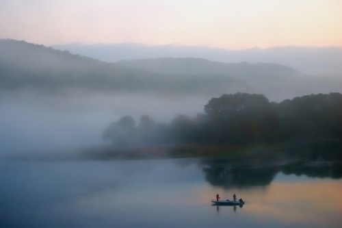 A serene lake at dawn, shrouded in mist, with two fishermen in a small boat silhouetted against the tranquil water.