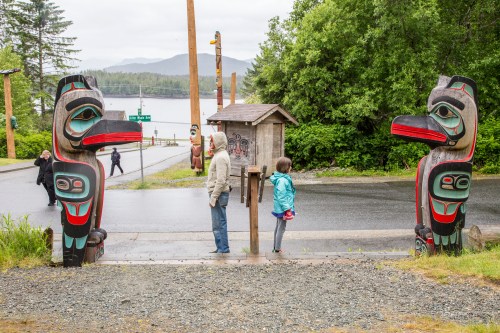 Two totem poles stand at a pathway, with a man and a child facing each other, surrounded by greenery and a waterfront view.