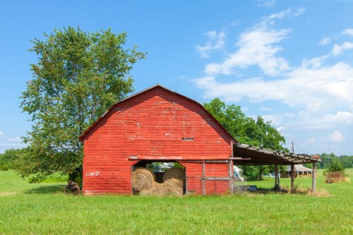 A red barn with a haystack, surrounded by green grass and trees under a blue sky with scattered clouds.