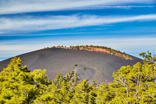 A volcanic landscape with a dark, sloping cone and green trees in the foreground under a blue sky with wispy clouds.