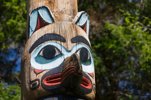 Close-up of a colorful totem pole featuring a carved animal face, surrounded by greenery and natural light.