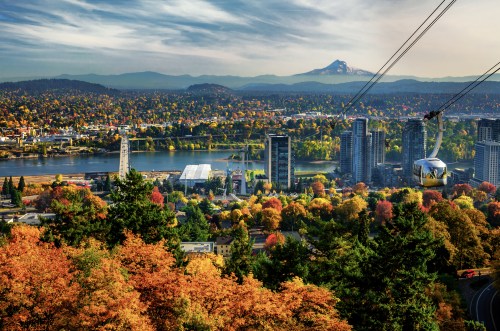 A scenic view of a city with autumn foliage, a river, and mountains in the background, featuring a cable car.