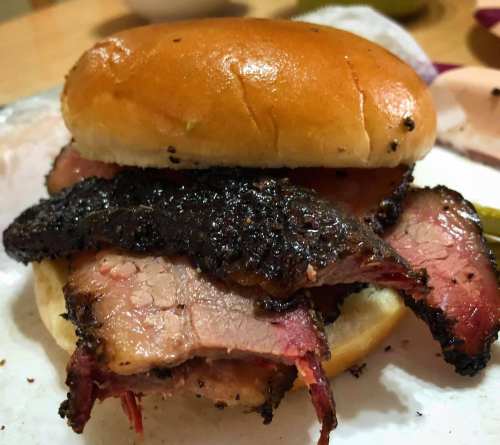A close-up of a sandwich with slices of smoked brisket on a soft bun, garnished with pickles.