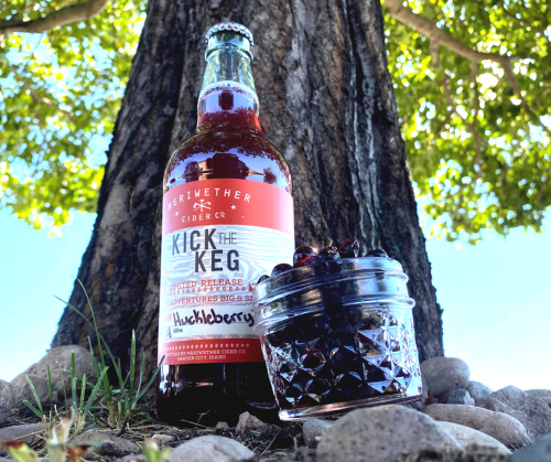 A bottle of huckleberry cider next to a jar of huckleberries, set against a tree and grassy ground.