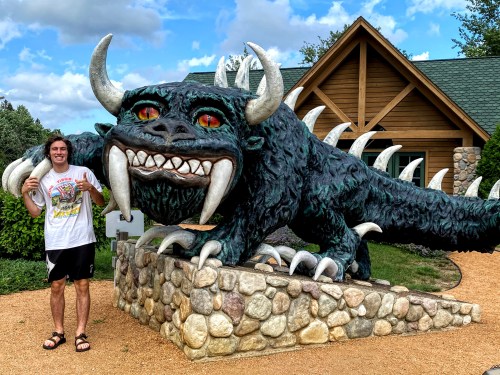 A person stands next to a large, colorful dragon sculpture with sharp teeth and bright eyes, in front of a wooden building.