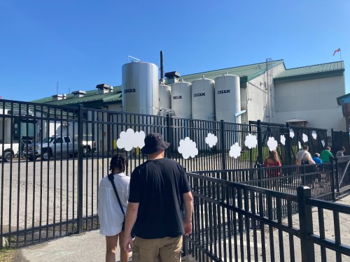 Two people walk towards a fenced area with large silos labeled "SUGAR," "MILK," and "CREAM" under a clear blue sky.