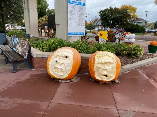 Two carved pumpkins with expressive faces, one showing fear and the other anger, placed on a paved area.