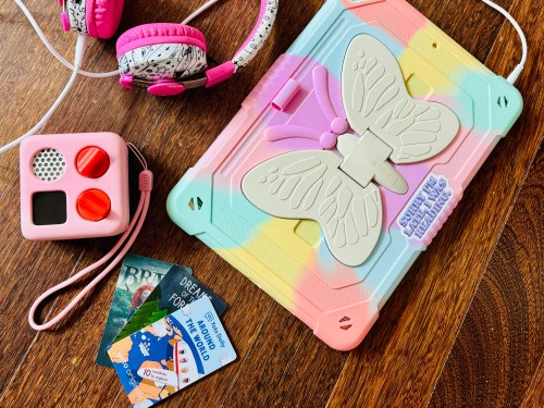 A colorful butterfly tablet case, pink headphones, a toy microphone, and travel-themed cards on a wooden surface.