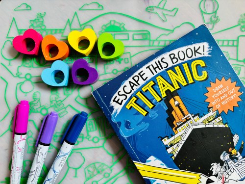 A colorful book titled "Escape This Book! Titanic" with markers and heart-shaped erasers on a playful background.