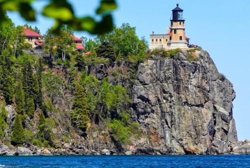 A lighthouse perched on a rocky cliff, surrounded by greenery and overlooking a calm blue lake.