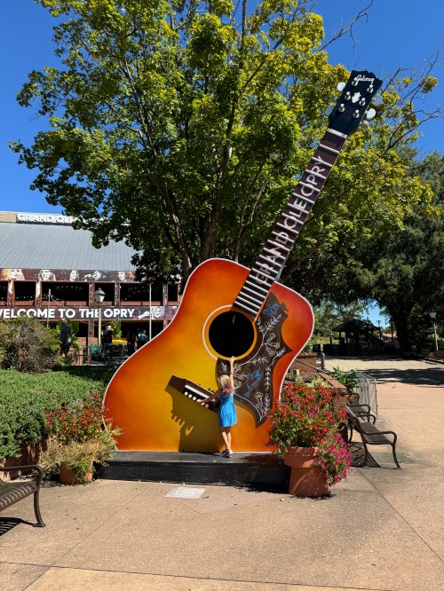 A child interacts with a large guitar sculpture outside the Grand Ole Opry, surrounded by trees and flowers.