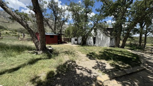 A rustic scene featuring a white house and a red barn surrounded by trees and grassy land under a blue sky.