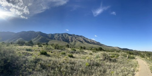 A panoramic view of mountains under a clear blue sky, with a path and green vegetation in the foreground.