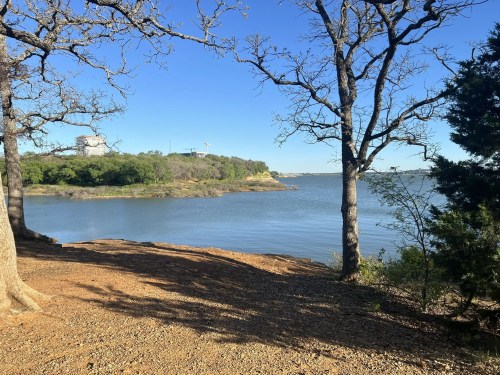 A serene lakeside view framed by trees, with calm water and a clear blue sky in the background.