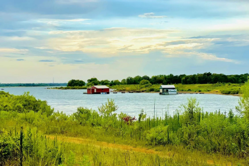 A serene landscape featuring a calm lake with two colorful houses on stilts, surrounded by greenery and a cloudy sky.