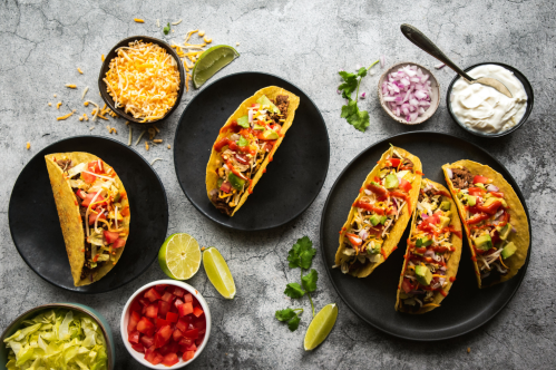 Three tacos filled with meat, cheese, and toppings, surrounded by bowls of ingredients like tomatoes, onions, and sour cream.