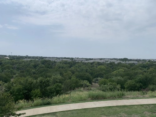 A panoramic view of a green landscape with trees and distant houses under a cloudy sky.